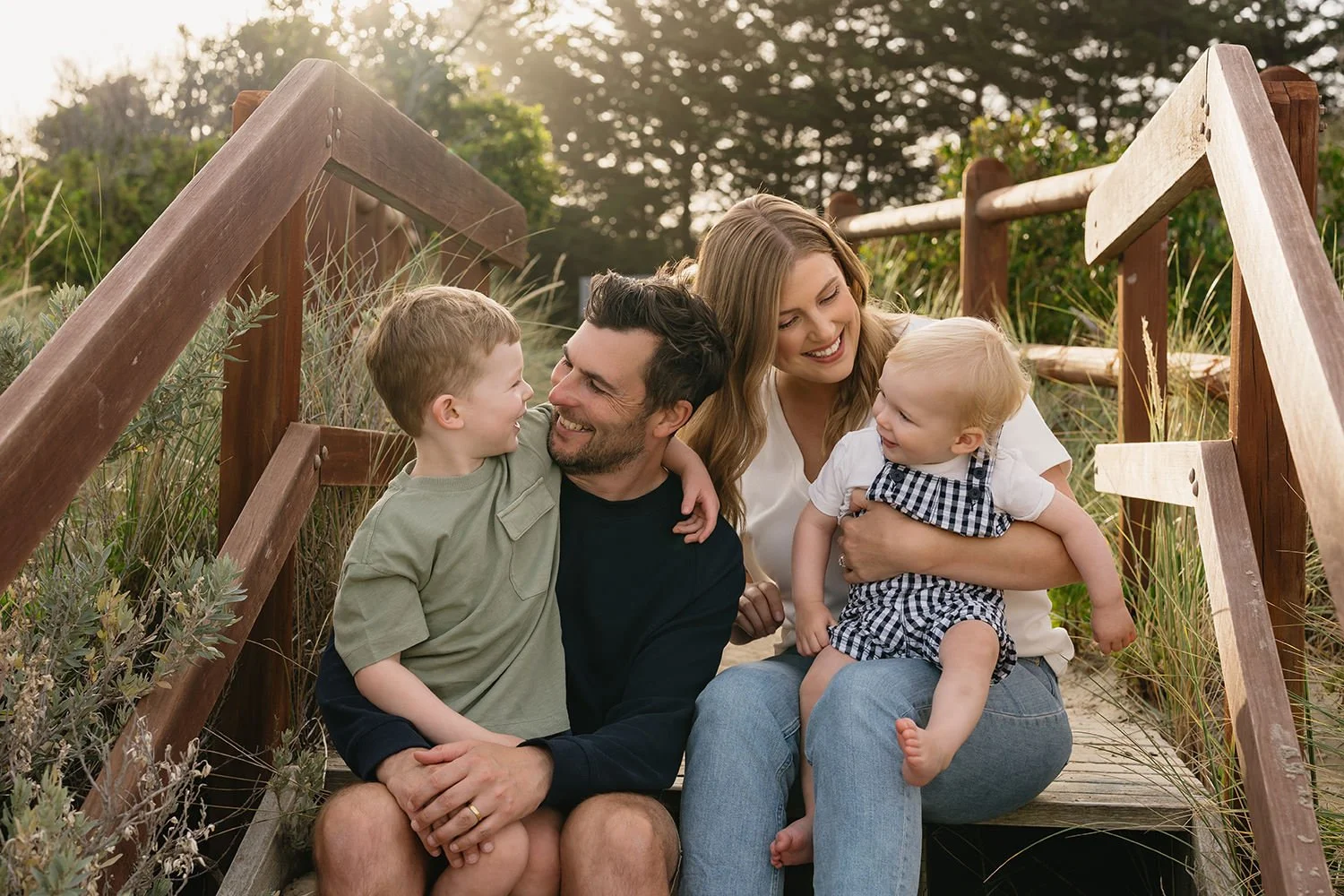 A family of four sitting on a wooden staircase outdoors, smiling and playing together in a natural setting with trees in the background.