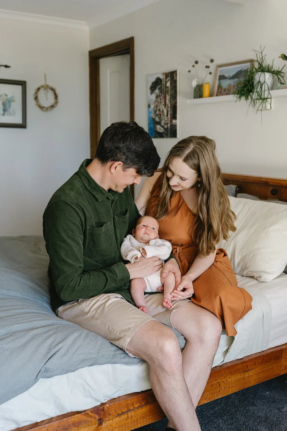 A happy family sitting on a bed, with a man holding a baby, and a woman leaning in, all smiling and looking at the baby.