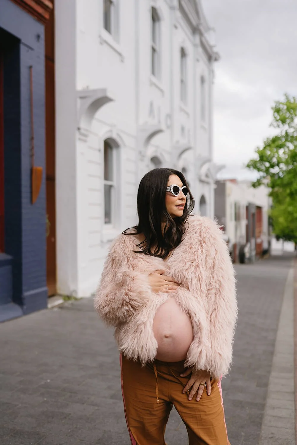 A pregnant woman with black hair and sunglasses standing outdoors on a city sidewalk, wearing a fluffy pink jacket and brown pants, with her hand on her belly and the other across her chest.
