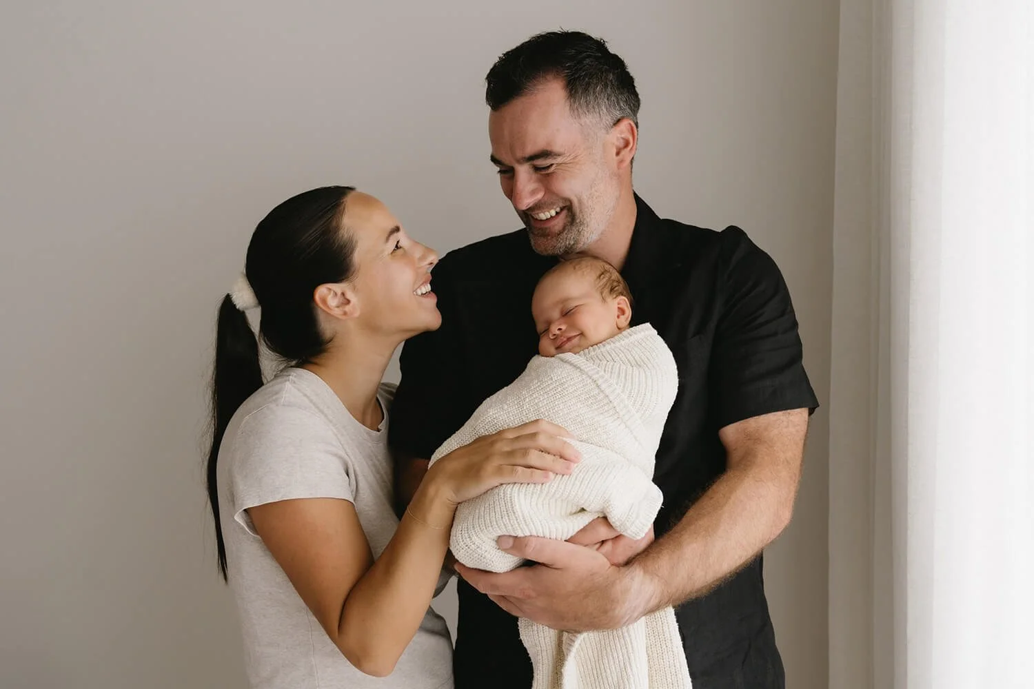 A happy family with a woman, man, and newborn baby smiling at each other indoors near a window.