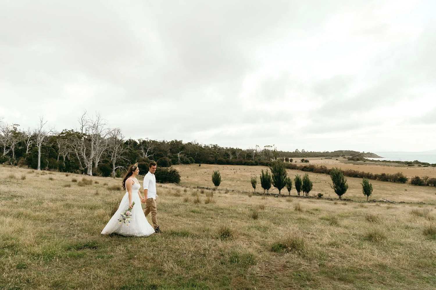 A bride and groom walking hand in hand across a grassy field with sparse trees, under a cloudy sky, in a rural landscape.
