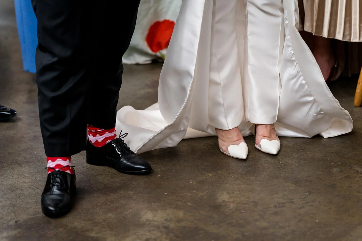 Close-up of a bride and groom's feet during a wedding ceremony, showing the bride in white heels with heart-shaped toes and the groom in black shoes with red and white striped socks, standing on a concrete floor.