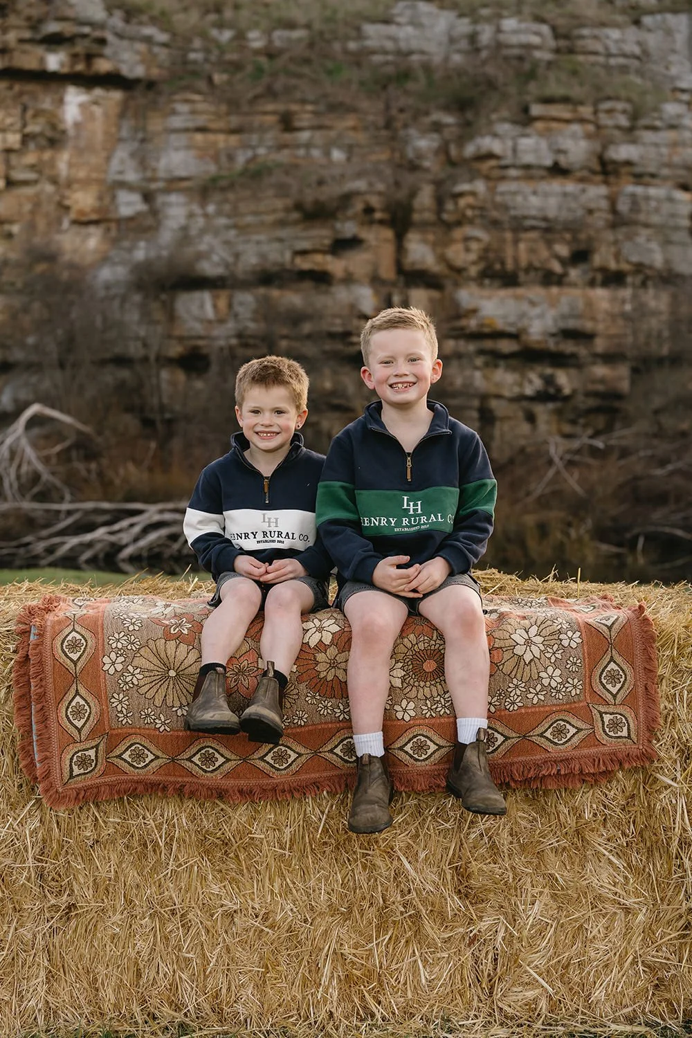 Two young boys sitting on a hay bale with a patterned rug, smiling, outdoors, with a rocky cliff and bare tree branches in the background.