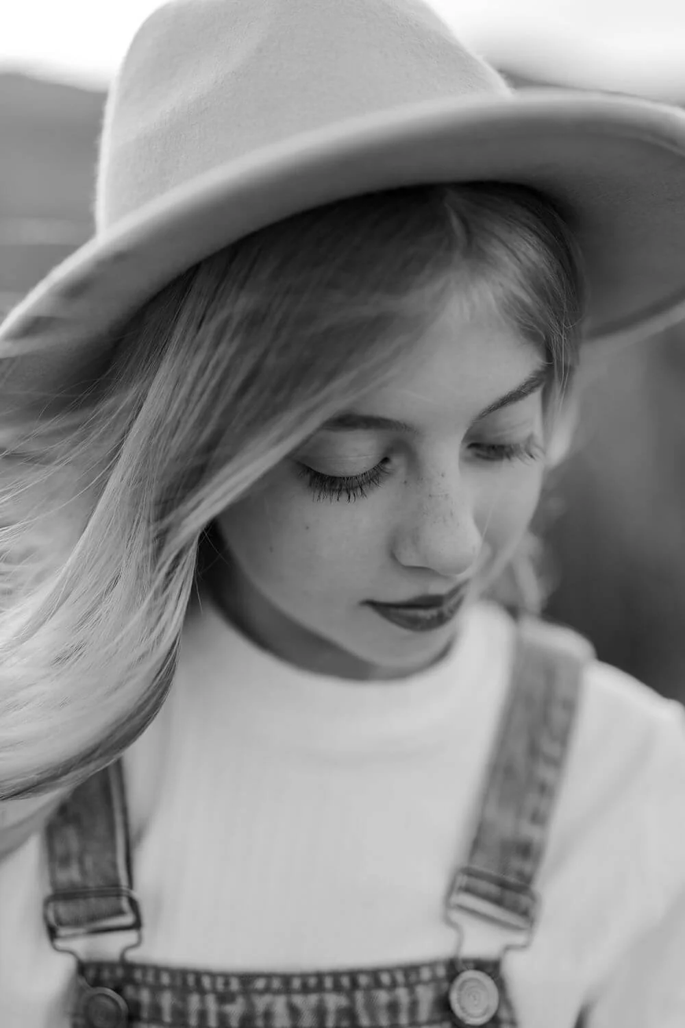 A young woman wearing a wide-brimmed hat, denim overalls, and a white shirt, with long, blonde hair and closed eyes.