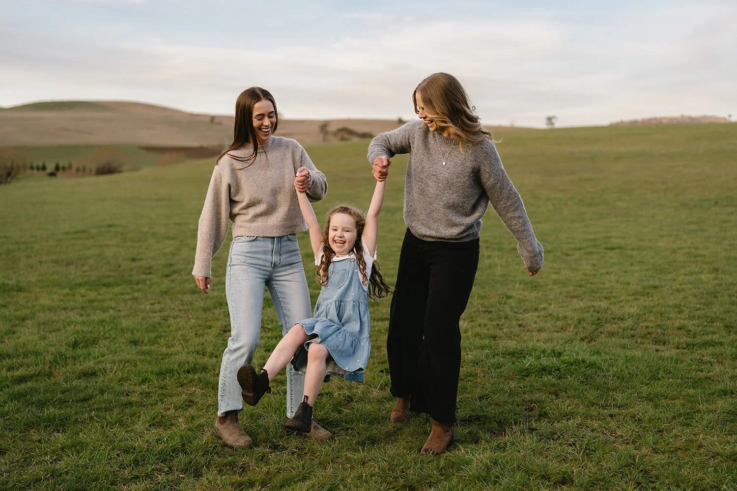 Two women and a young girl smiling and holding hands while walking on a grassy field. The girl is sitting on a swing being pulled by the women. The background shows rolling hills under a cloudy sky.