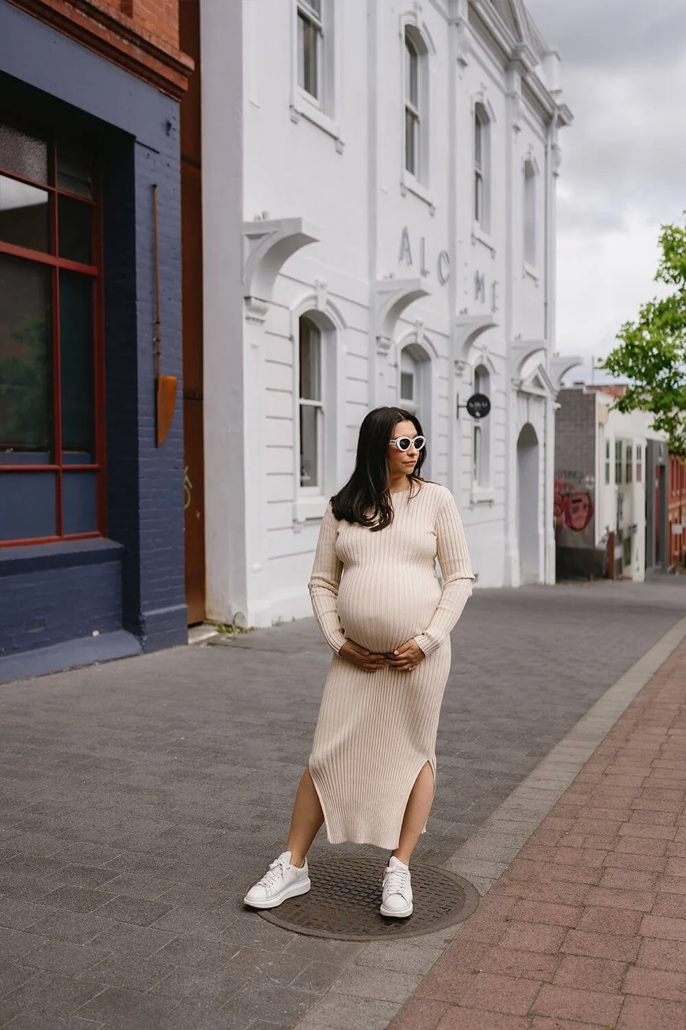 Pregnant woman wearing beige ribbed dress and white sneakers standing on a sidewalk in front of white and blue buildings, holding her belly with both hands and wearing sunglasses.