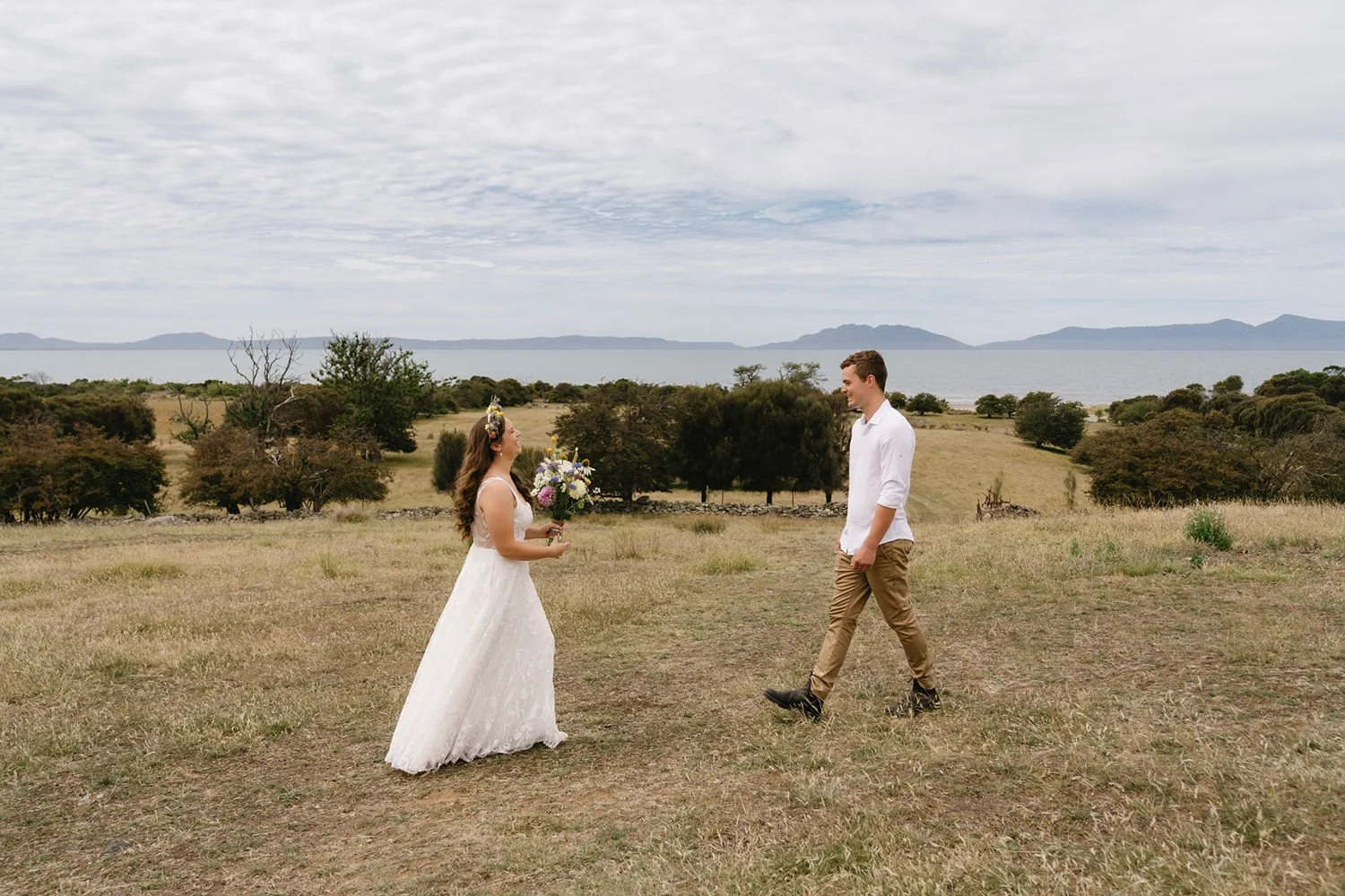 A bride in a white wedding dress holding a bouquet standing on a grassy field, smiling at a groom in a white shirt and tan pants, near a scenic waterfront landscape with trees, hills, and mountains in the background under a partly cloudy sky.