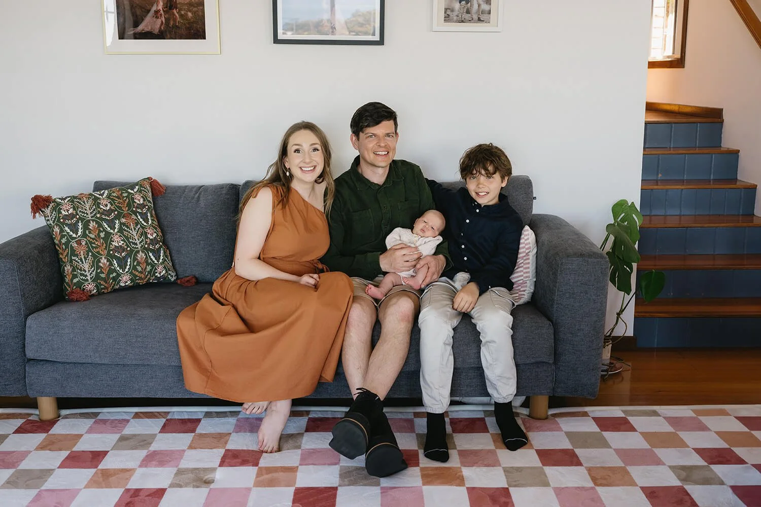 A happy family sitting together on a gray couch in a living room, includes a woman, a man holding a baby, and a boy, with decorative pillows and a plant nearby.