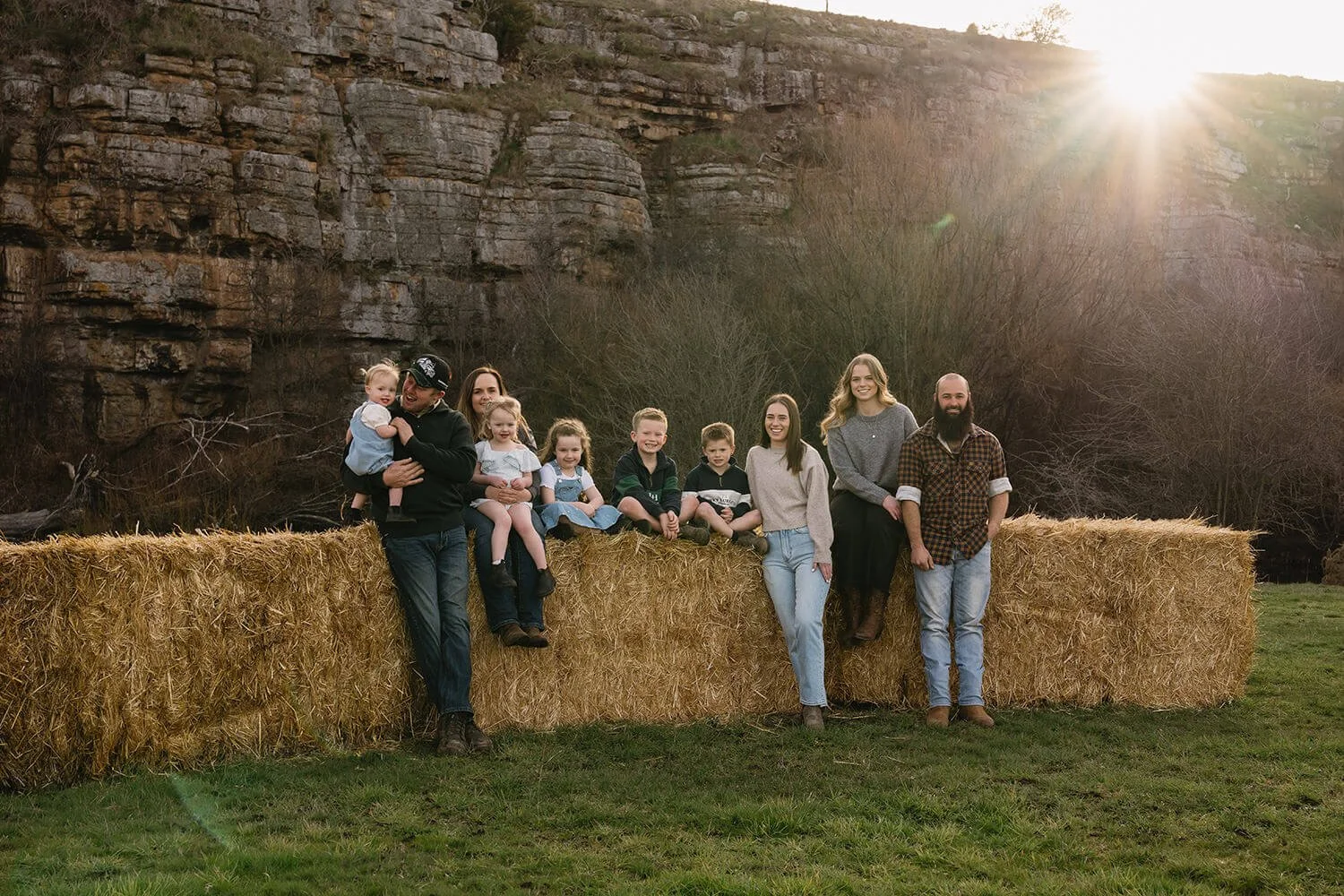 A large family gathered outdoors on a hay bale with a rocky hill and trees in the background, during sunset.