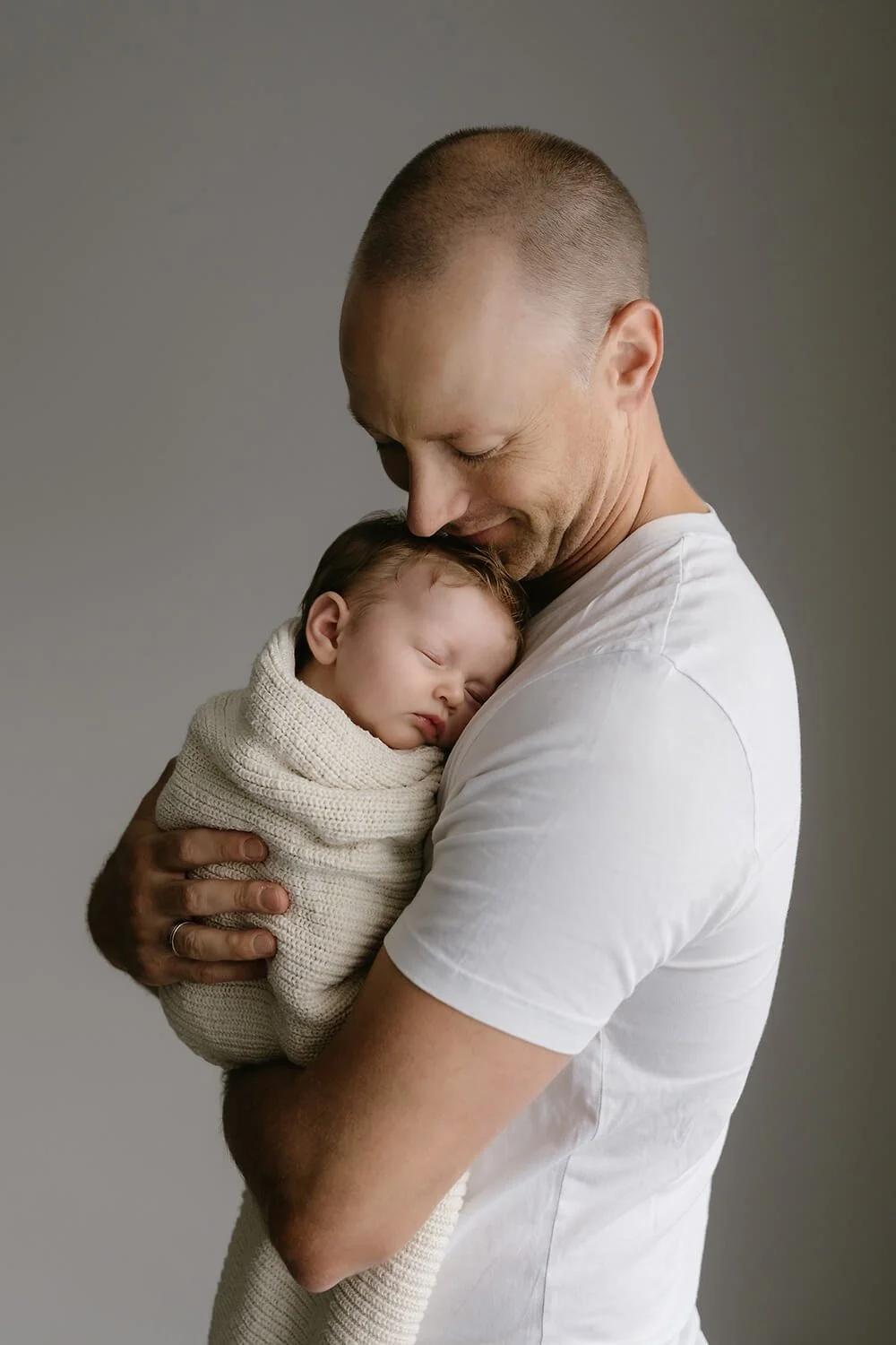 A man holding a sleeping baby wrapped in a knitted blanket, close to his chest in a tender, loving pose.