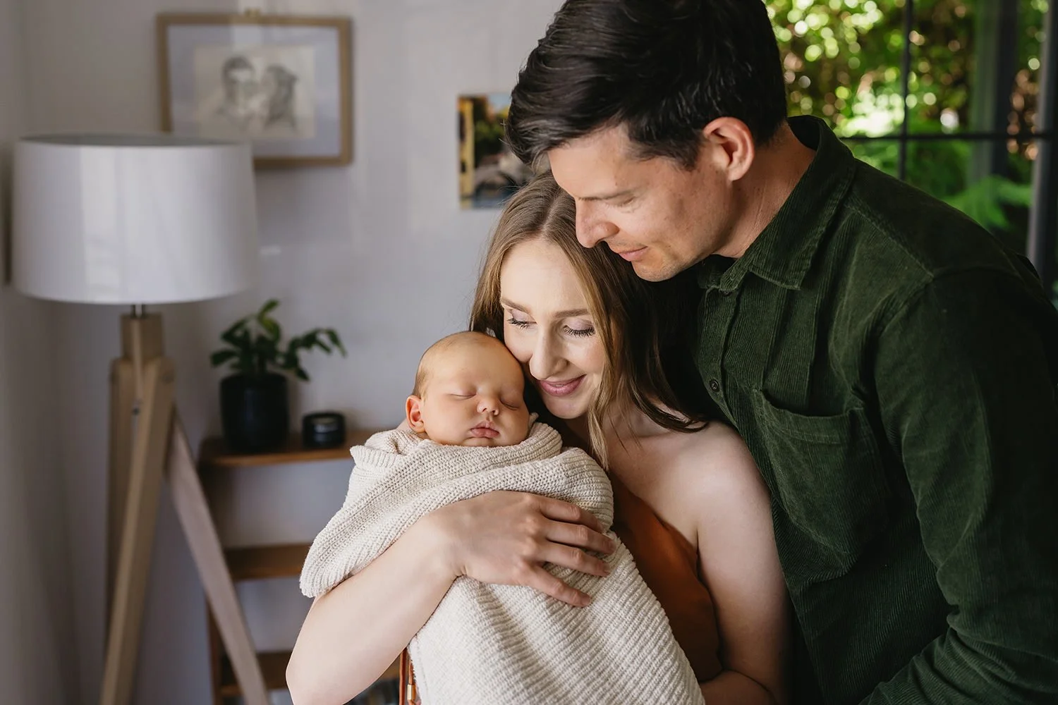 A family of three, a woman, a man, and a sleeping baby, cuddling indoors with a window showing greenery outside.