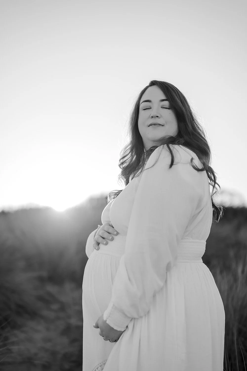 Black and white photograph of a pregnant woman standing outdoors, her eyes closed and head slightly tilted, with a serene expression on her face. She is wearing a long, flowing dress and has long, dark hair. The background features a blurry natural l