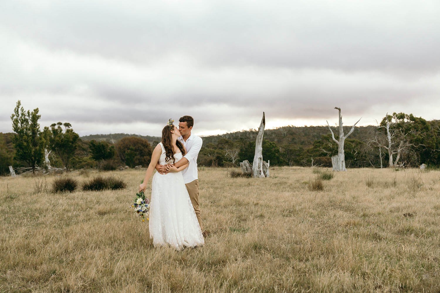 A couple in wedding attire sharing a kiss in a grassy field with dry trees and cloudy sky in the background.