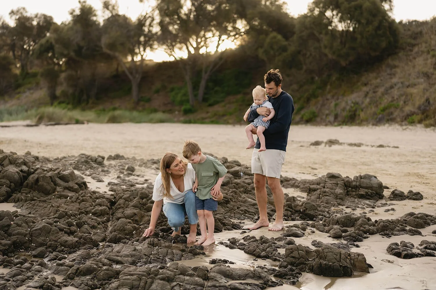 A family of four on a rocky beach during sunset. The mother kneels and looks at her young son, who is standing on the rocks, while the father holds a baby girl in his arms. The background features sandy shore, trees, and a sunset sky.