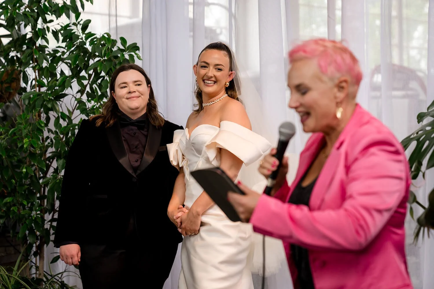 Three women at a wedding, with one woman in a white wedding dress smiling, another woman in black attire standing next to her, and a woman in pink attire holding a microphone and reading from a tablet, with plants and sheer curtains in the background