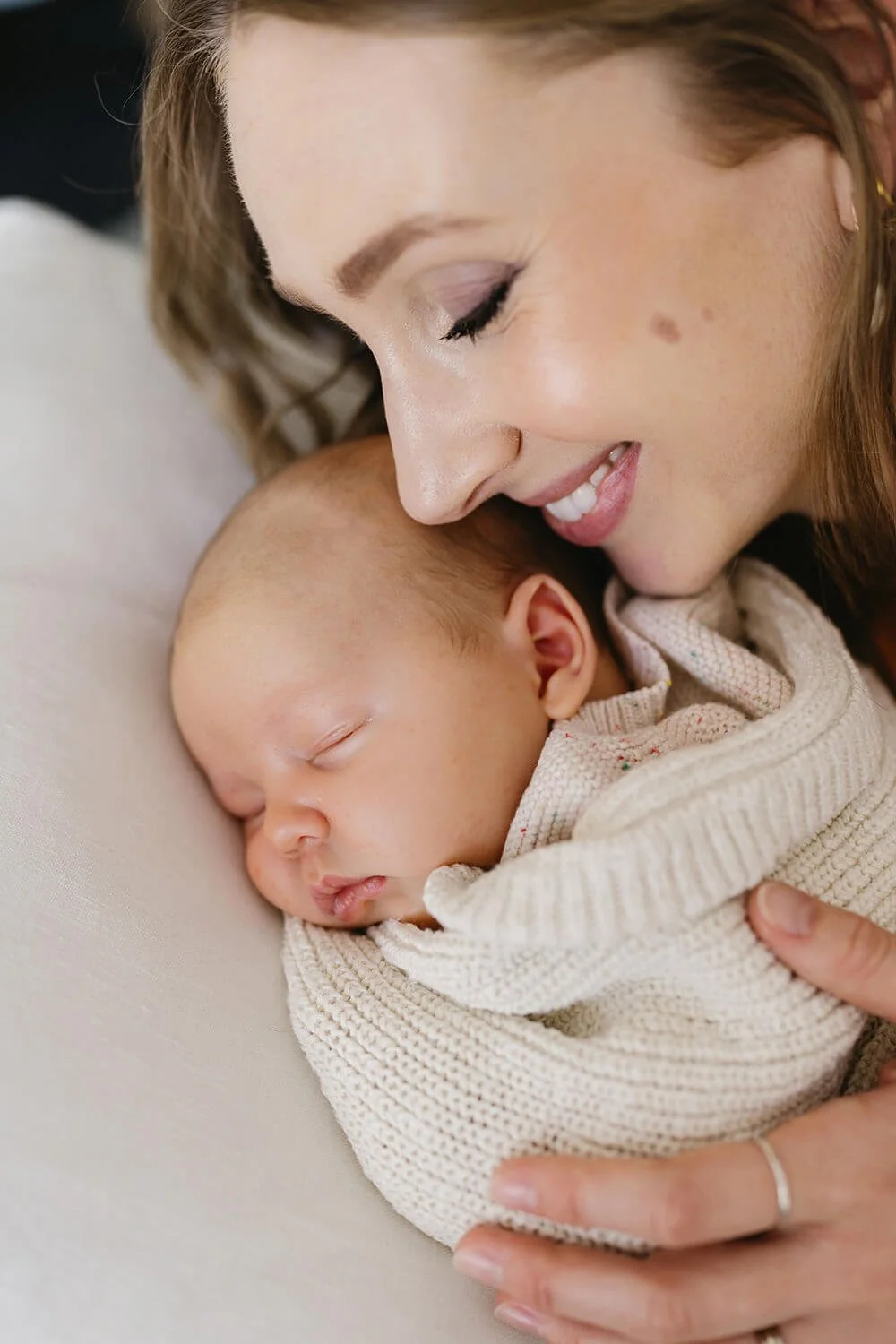 A woman is holding a sleeping baby wrapped in a beige knitted blanket, close to her face, smiling gently.