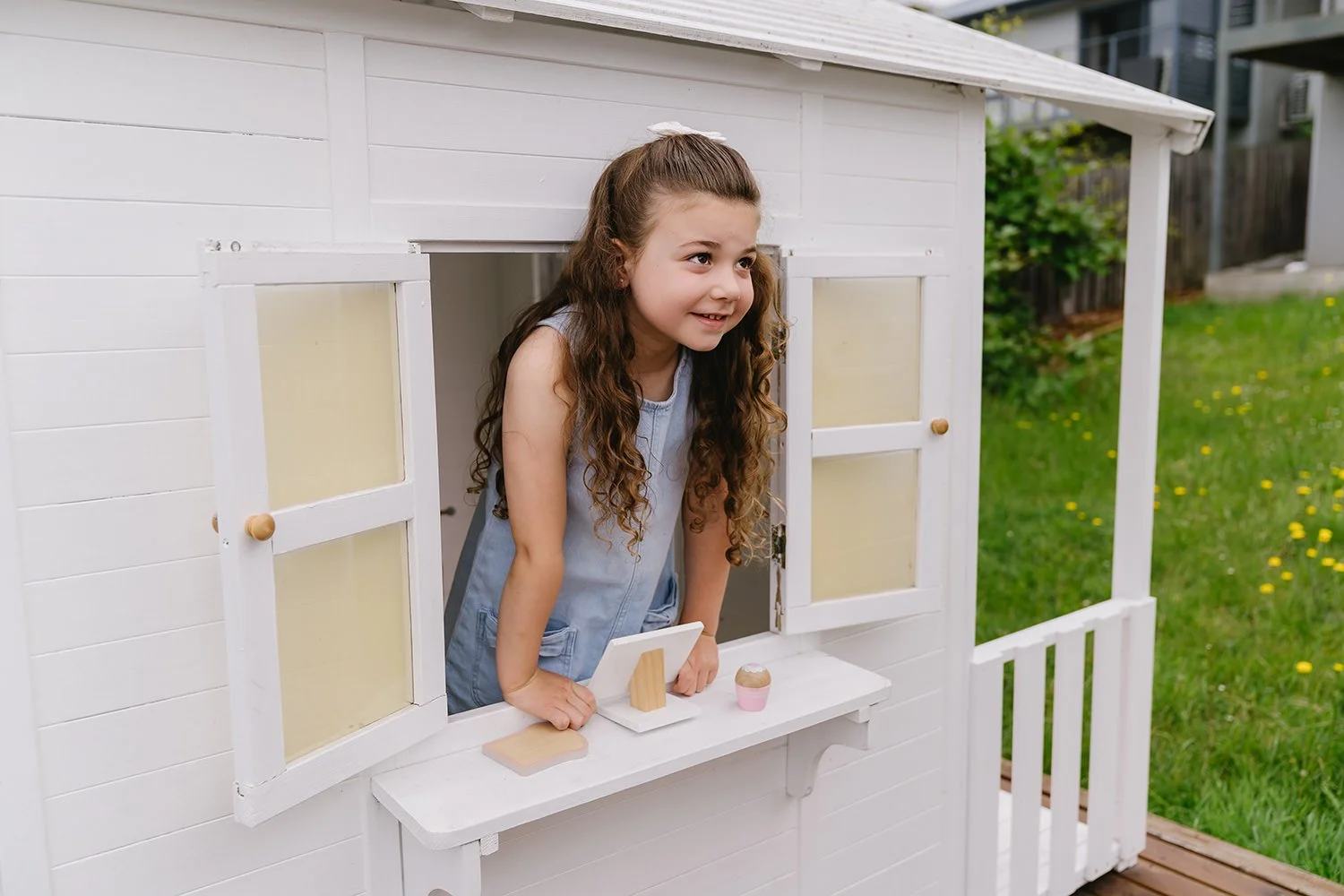 A girl with long curly hair leaning out of a white playhouse window, smiling, with a small white counter holding a miniature mirror and a cupcake on it, in a grassy backyard.