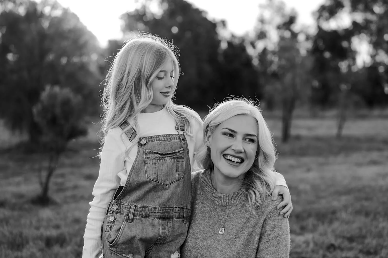 A black and white photo of a girl with long hair and a woman with short, wavy hair in an outdoor park setting, smiling and looking at each other.