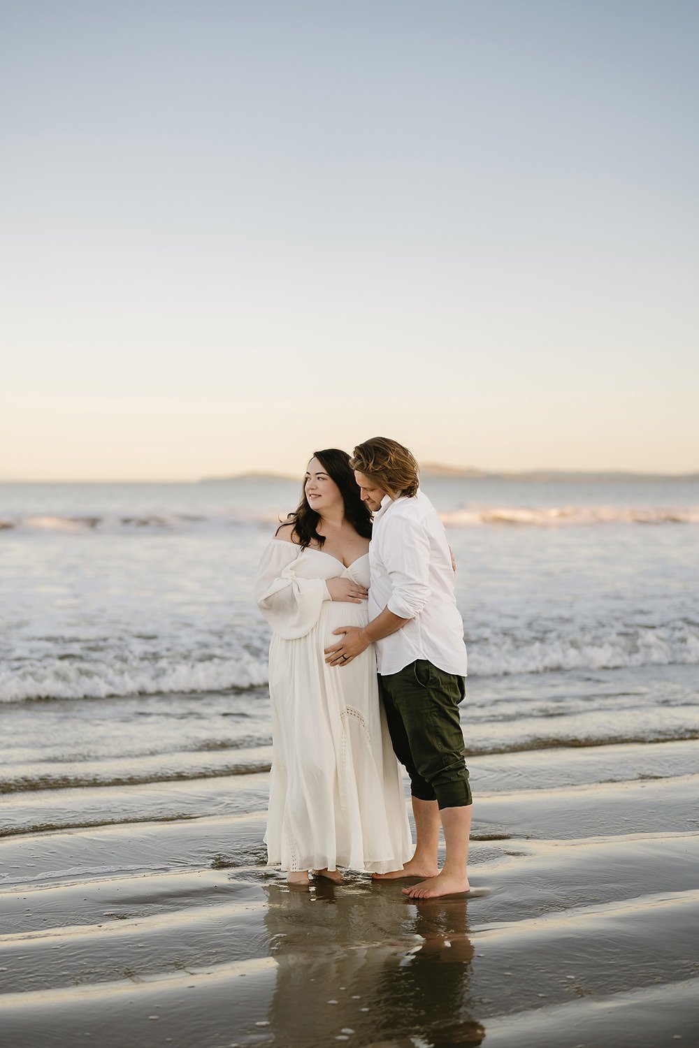 A pregnant woman and a man standing barefoot in the sand at the beach, holding each other and looking at each other during sunset.