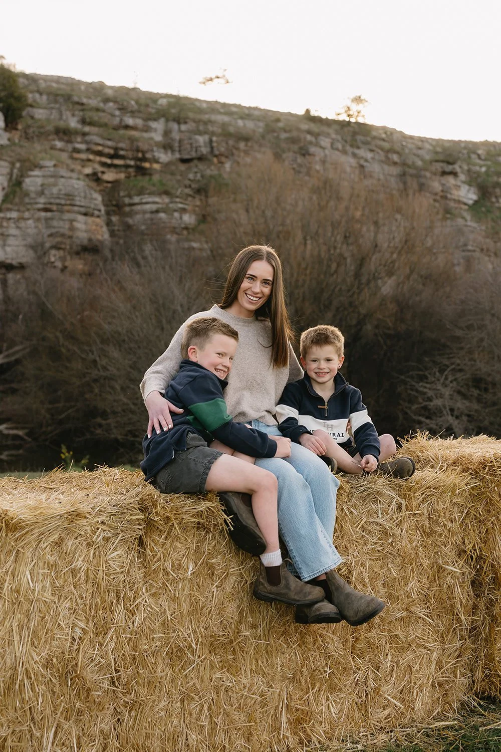 A woman and two young boys sitting on a hay bale outdoors, smiling at the camera, with rocky hills and trees in the background during daytime.