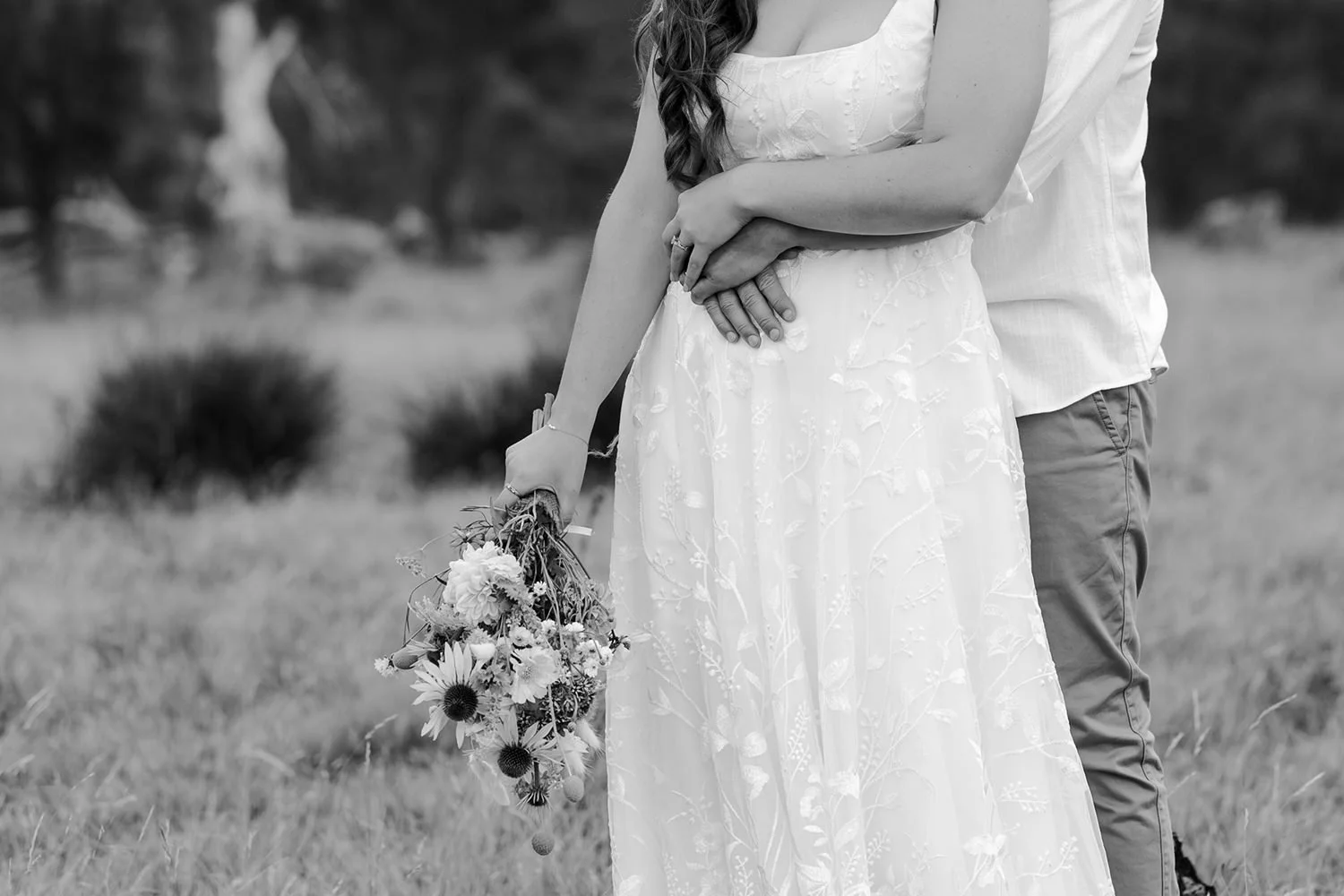 A couple standing outdoors: a woman in a white floral dress holding a bouquet of flowers, and a man standing behind her with his arm around her waist.