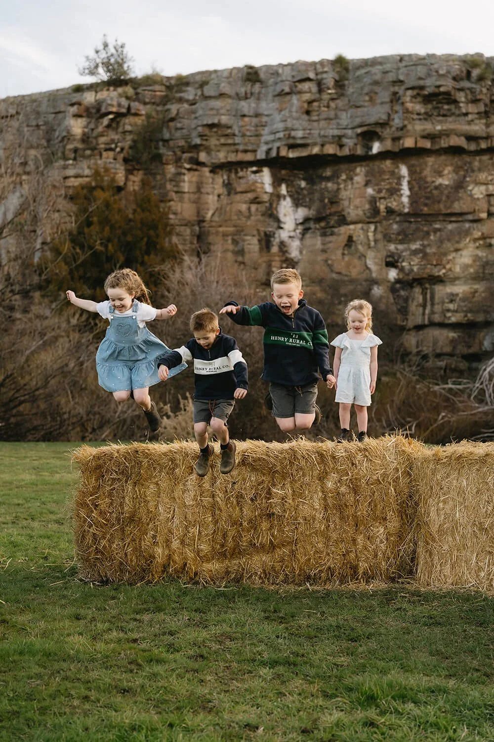 Four children jumping off a hay bale in a field with a rocky cliff and sparse trees in the background.