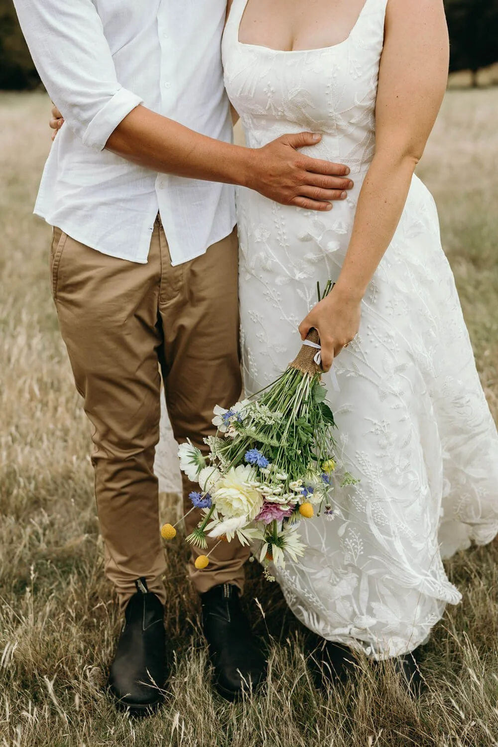 A couple standing outdoors; the woman wears a white lace dress and holds a bouquet of mixed flowers, while the man wears a white shirt and tan pants.