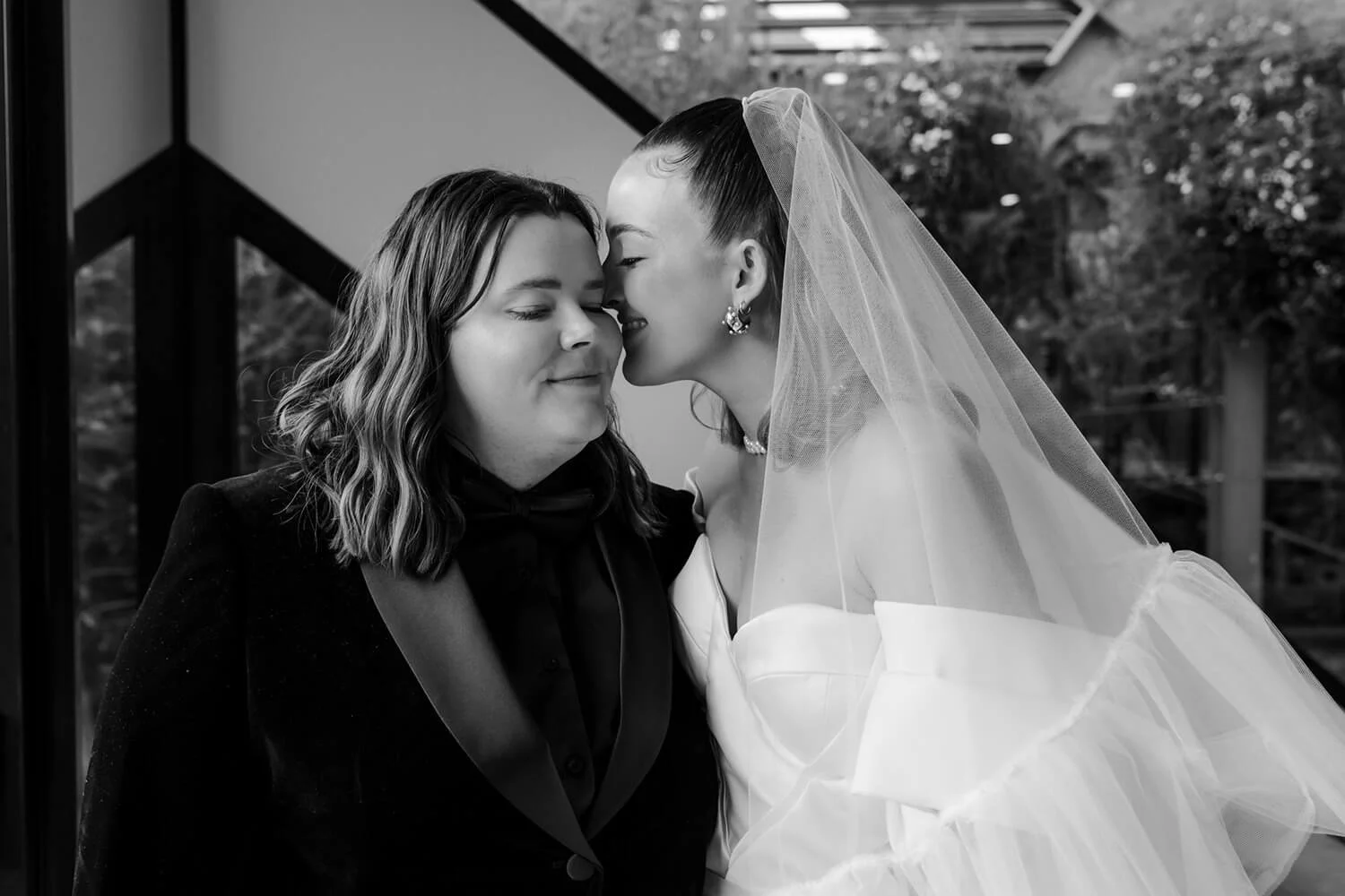 Two women sharing a tender moment, one in a wedding dress and veil, the other in a dark suit, nose to nose with eyes closed, smiling gently.