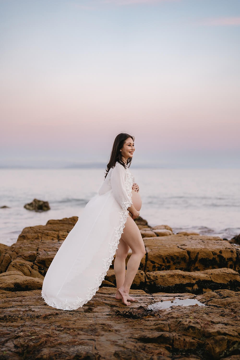 A pregnant woman standing on rocks at the beach during sunset, smiling, wearing a white lace robe.