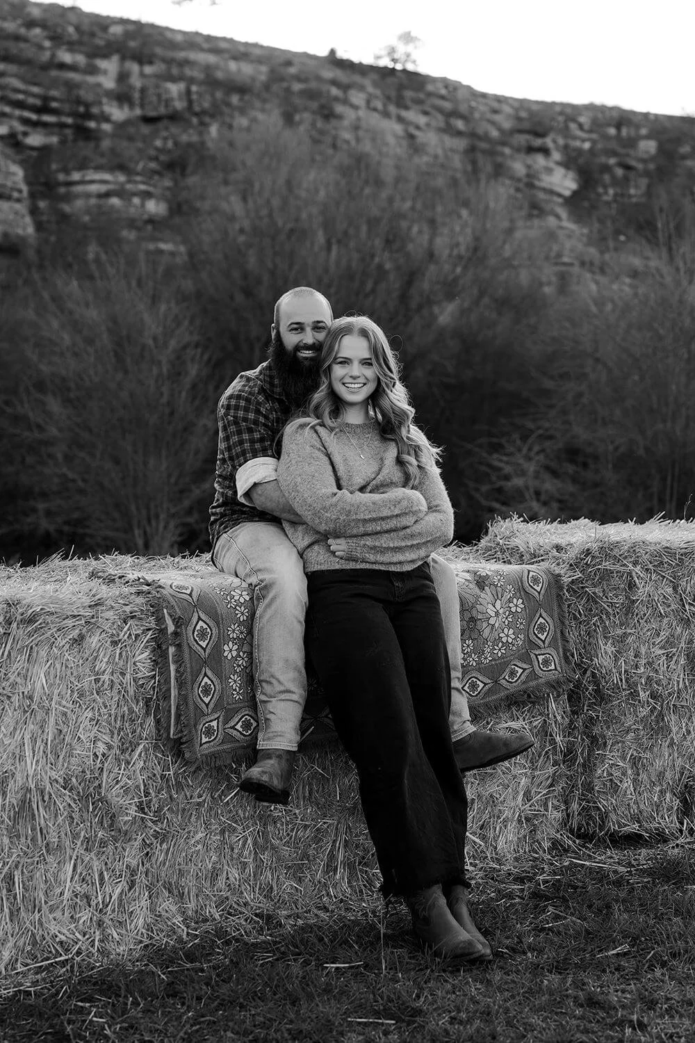 A black-and-white photo of a smiling couple sitting on hay bales outdoors, with a wooded hillside in the background.