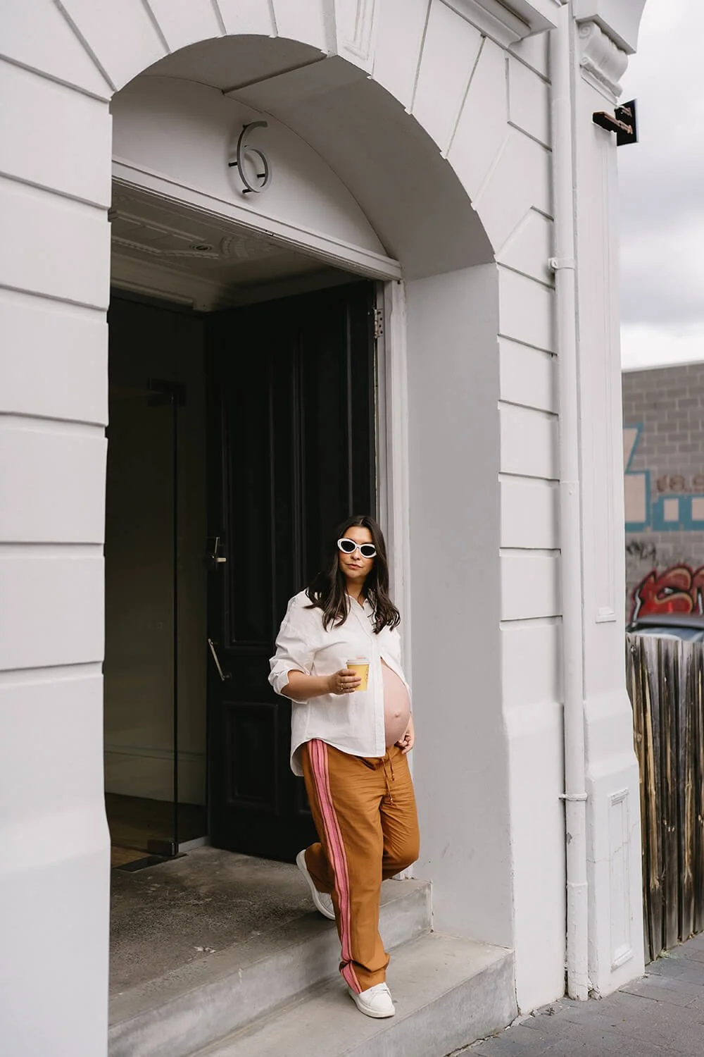 Pregnant woman with dark hair, wearing sunglasses, a white shirt, beige pants with pink stripes, and white sneakers, standing on a concrete step outside a building, holding a yellow cup.