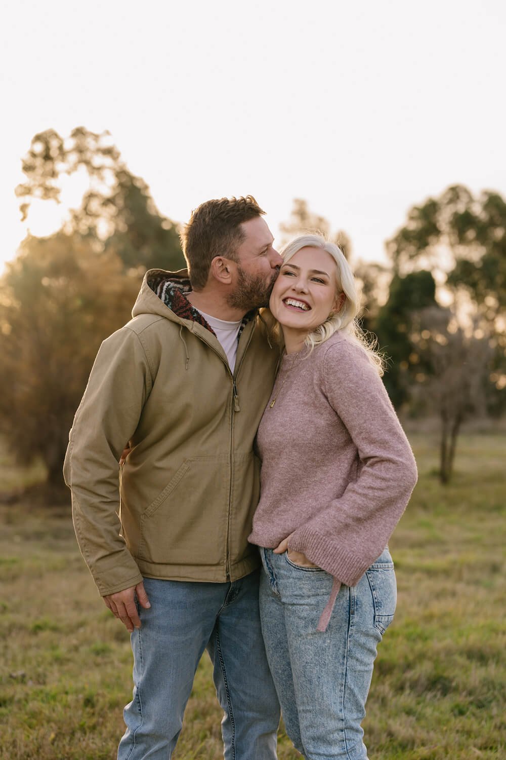 A man kisses a laughing woman on the cheek outdoors during sunset, wearing casual clothes in a grassy field with trees in the background.