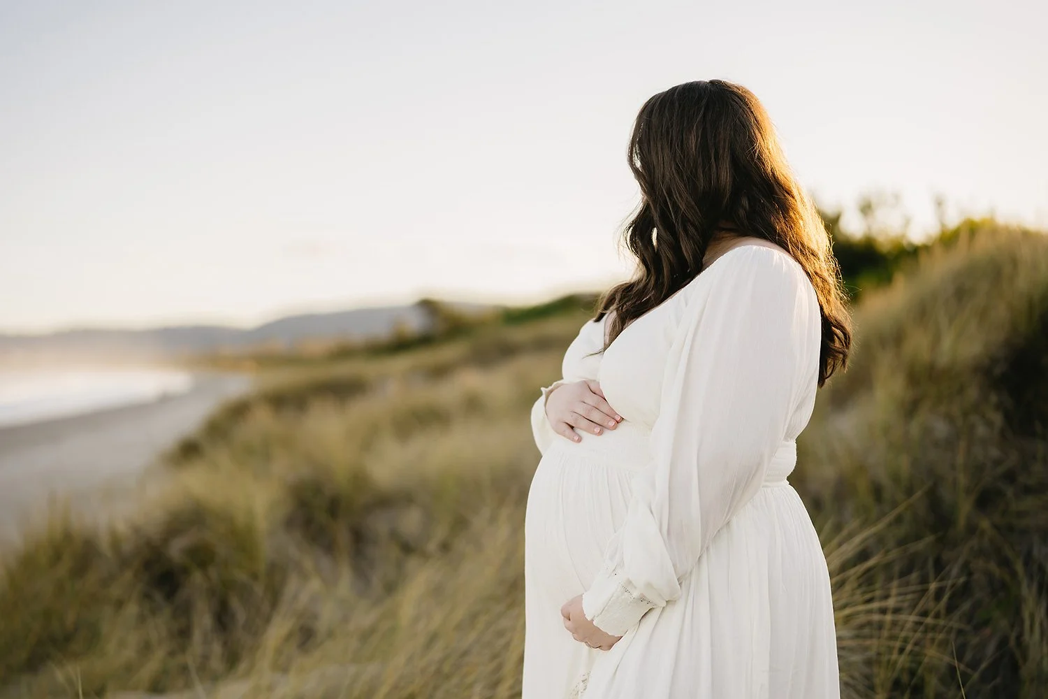 A pregnant woman with long brown hair wearing a white dress, standing on a beach near tall grasses during sunset, looking to the side with arms crossed over her belly.
