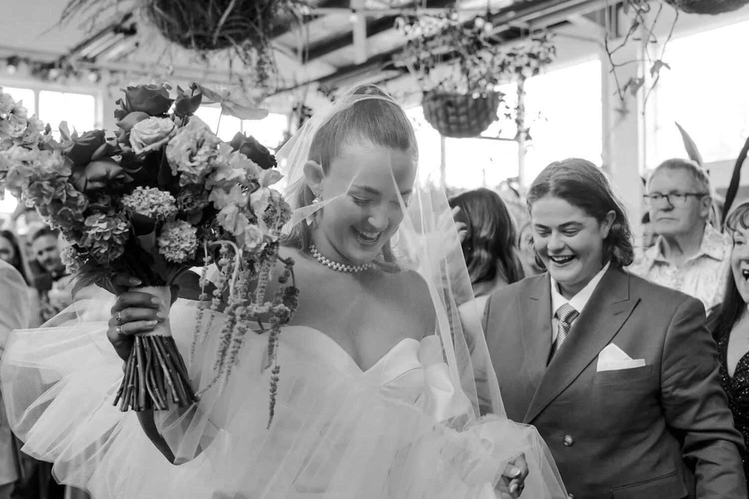 A joyful bride in a wedding dress holding a large bouquet of flowers, surrounded by wedding guests in a bright, indoor venue.