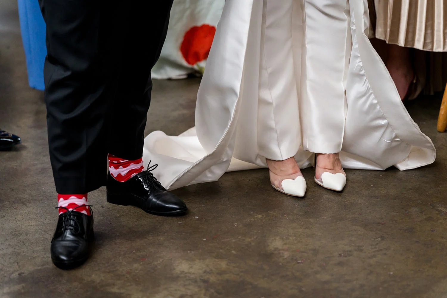 Close-up of a bride's feet in white wedding shoes on a wedding dress train, with guests' feet visible nearby.