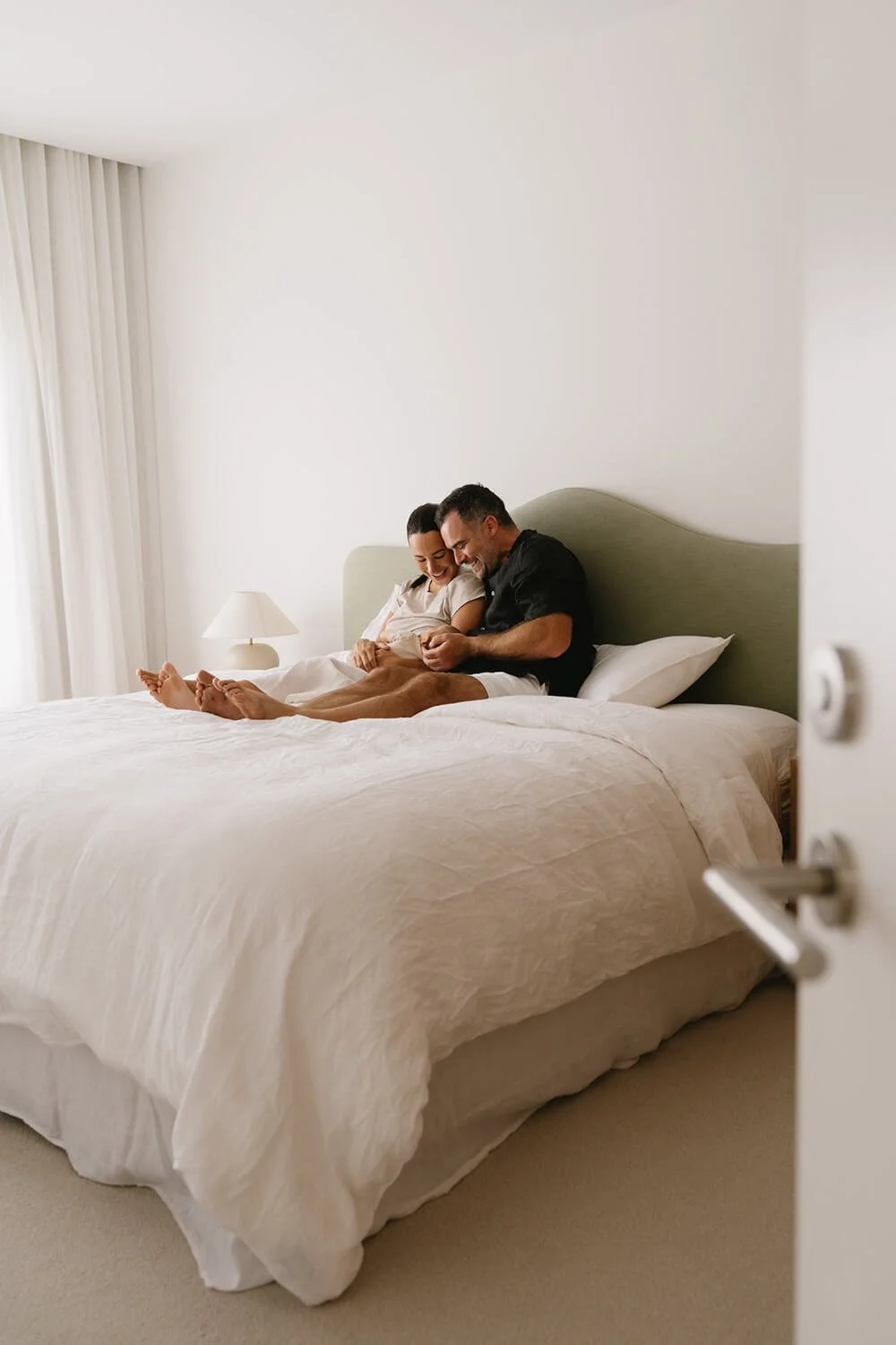 A couple sitting on a bed, smiling and looking at a phone, in a bright, minimal bedroom with white bedding, a bedside lamp, and curtains.