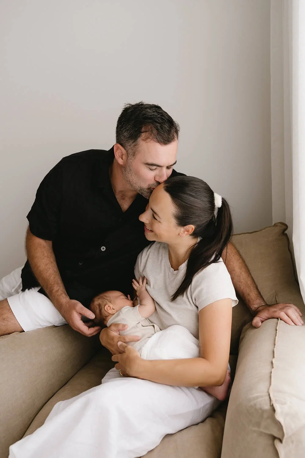 A family of three parenting a newborn on a beige sofa in a cozy, softly lit room.