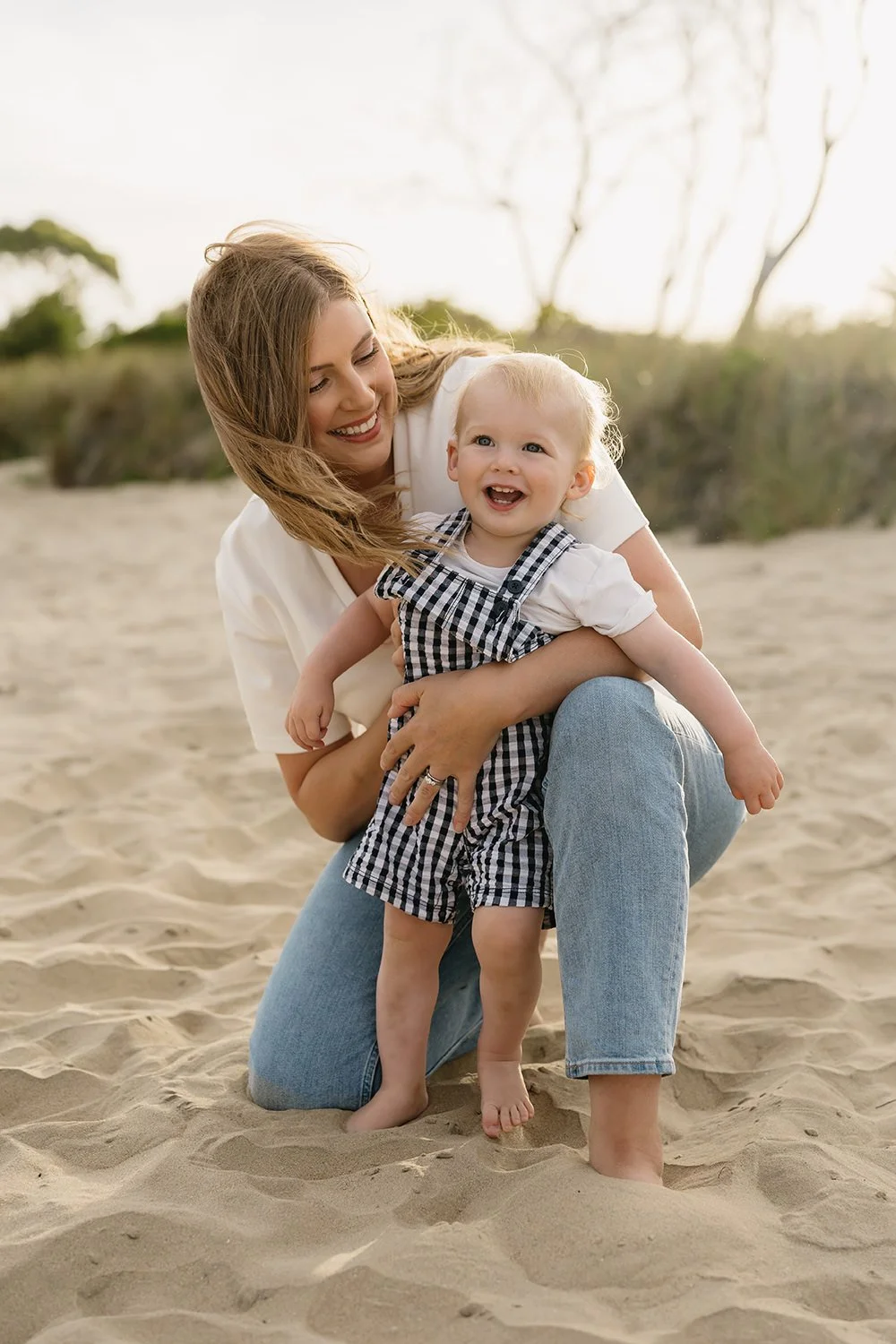 A young woman and a toddler playing together on the sand at the beach, smiling and enjoying the moment.