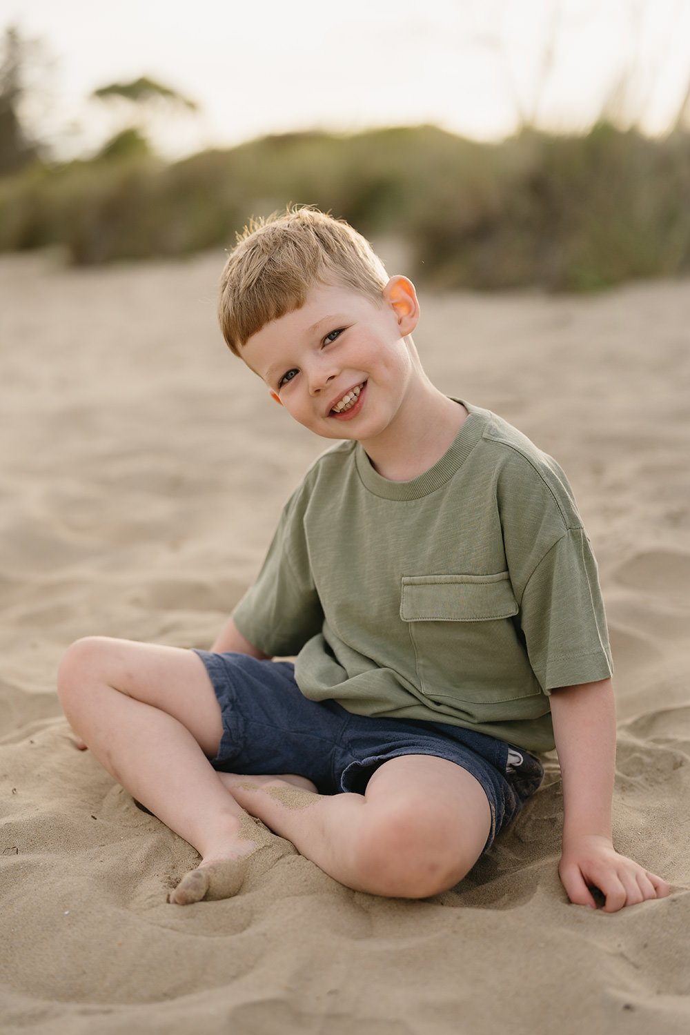A young boy with light brown hair, wearing a green t-shirt and dark blue shorts, sitting on sandy ground at the beach, smiling and looking at the camera.