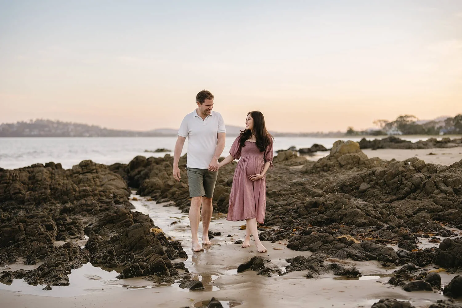 A couple walking hand in hand on a rocky beach at sunset, the woman cradling her pregnant belly, both smiling and looking at each other.