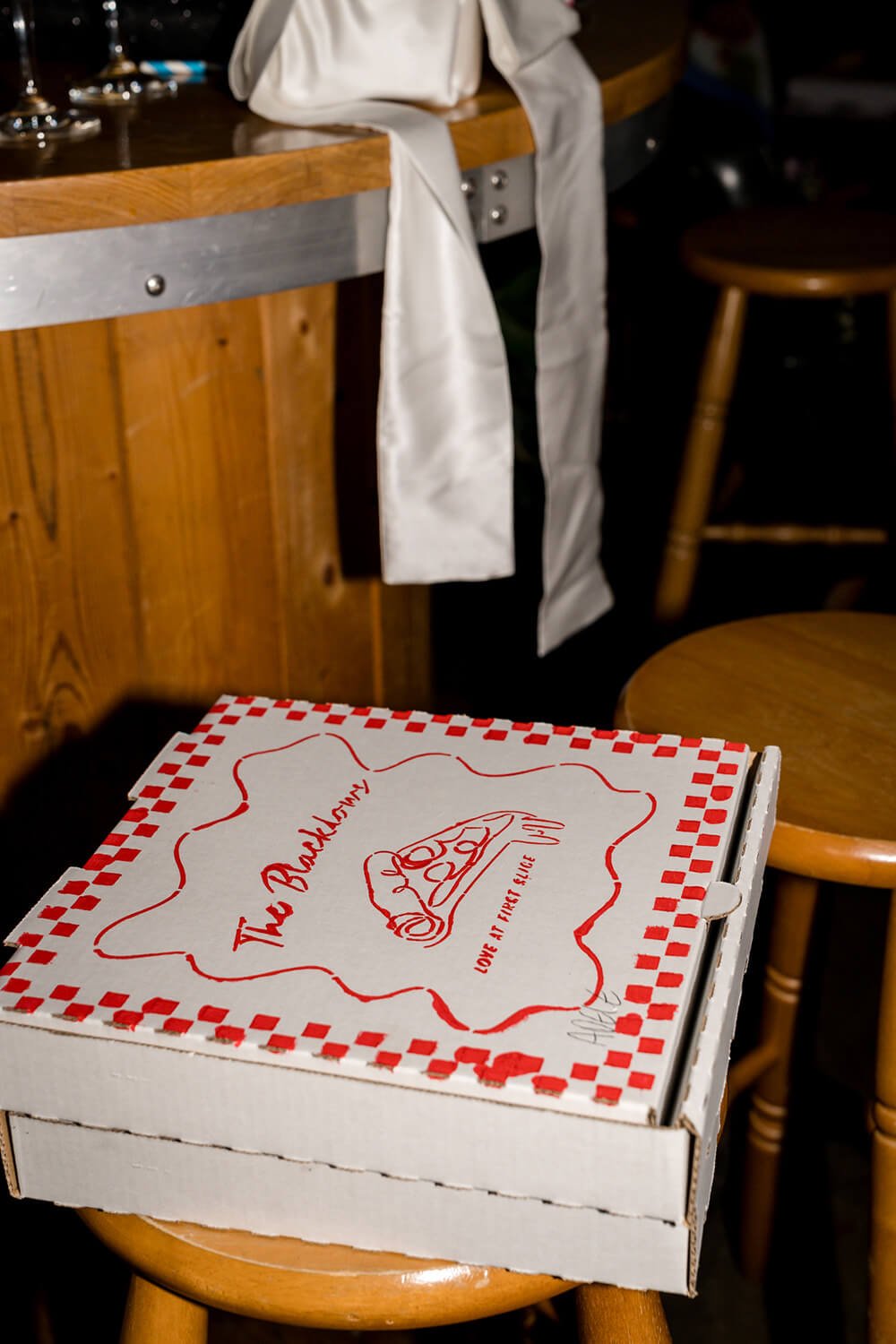 A pizza box with a red and white checkered border on a small wooden stool in a restaurant, with a rotary phone sketch and text that says "The Bricklayer" and "Love at first slice" on the pizza box. In the background, a white cloth napkin hangs over a