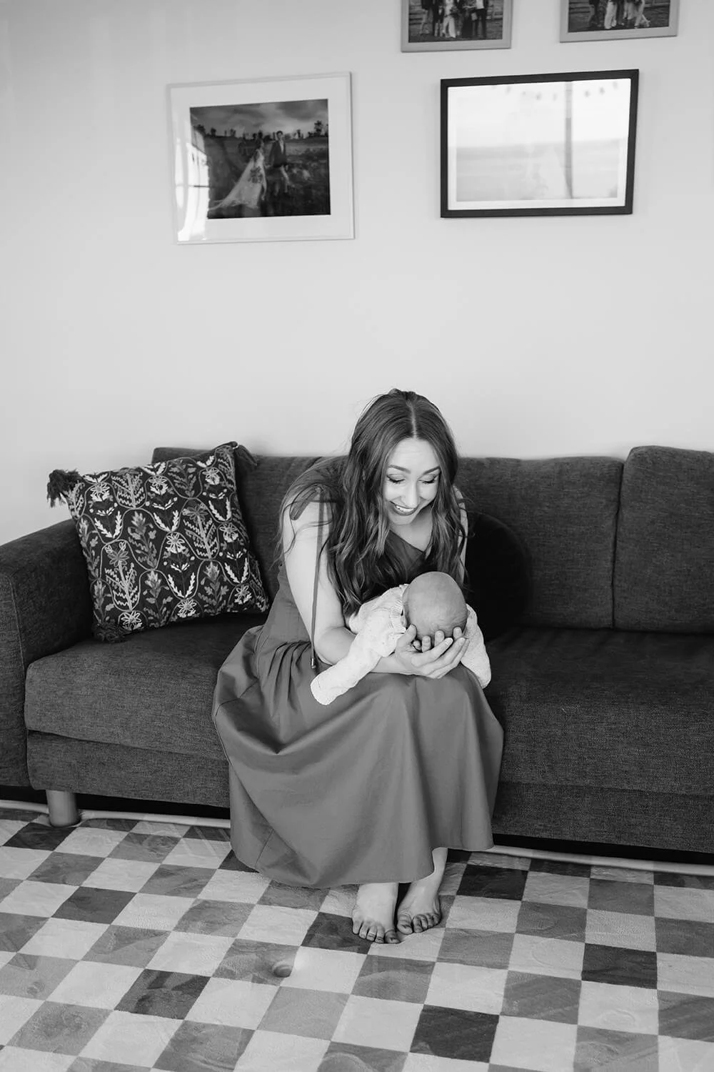 A woman sitting on a sofa, holding a newborn baby, smiling and looking at the baby, in a living room with framed photos on the wall and a patterned pillow on the sofa.