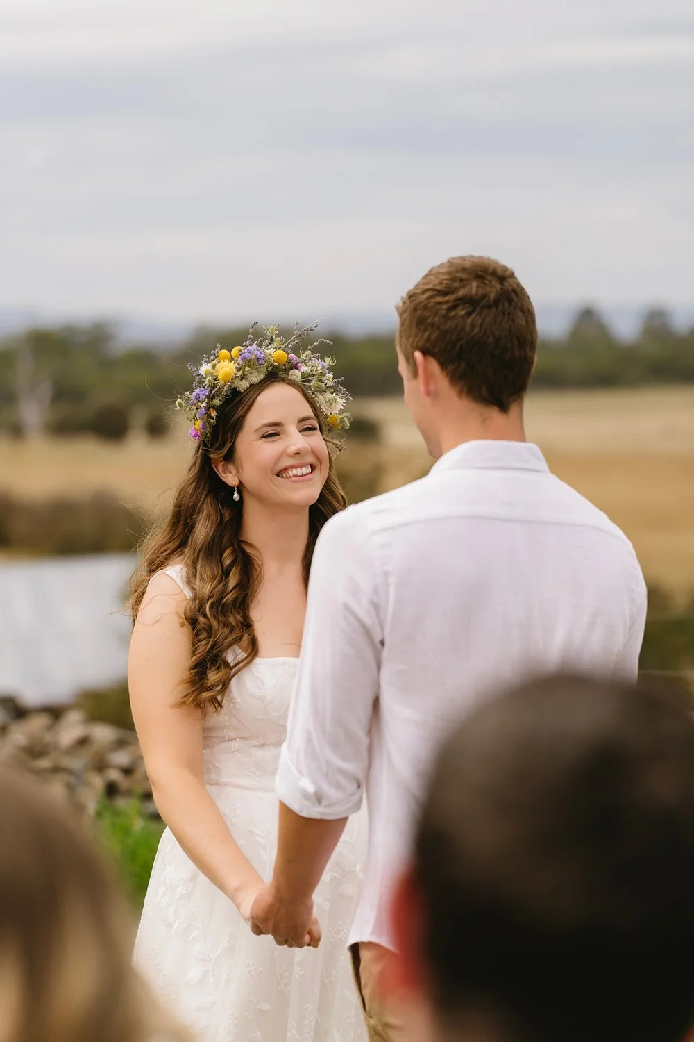 A smiling bride with long wavy hair wearing a flower crown and a white dress, holding hands with a groom in a white shirt during an outdoor wedding ceremony.