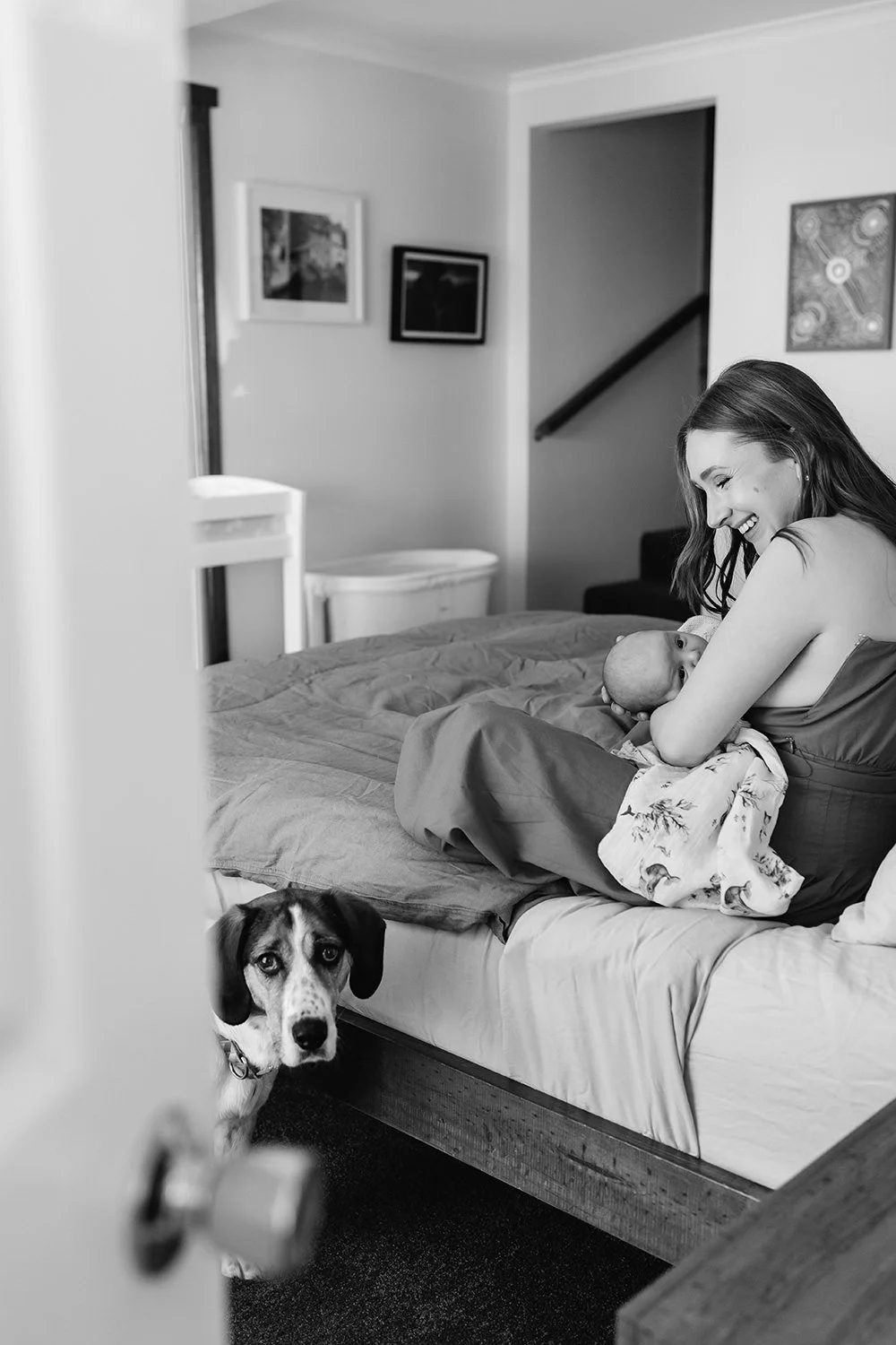 A woman holding a newborn baby on a bed, smiling, with a dog looking out from behind the bed in a cozy room.