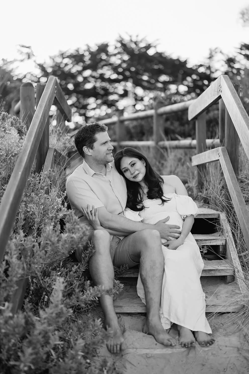 A black and white photo of a couple sitting on outdoor wooden stairs, with the woman visibly pregnant. The man is smiling and looking at the woman, who is resting her head on his shoulder. They are surrounded by plants and trees.