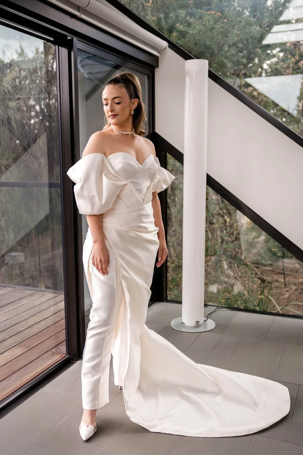 Woman in a white wedding dress with off-shoulder puff sleeves standing indoors near glass doors, with a neutral expression and her eyes closed.