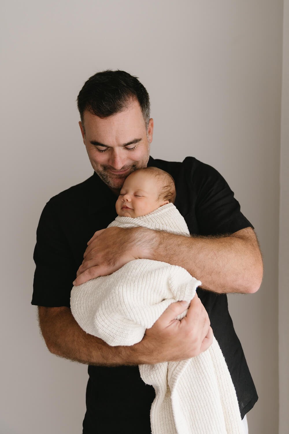 A man holding a sleeping baby wrapped in a white blanket, both smiling gently, against a plain neutral background.