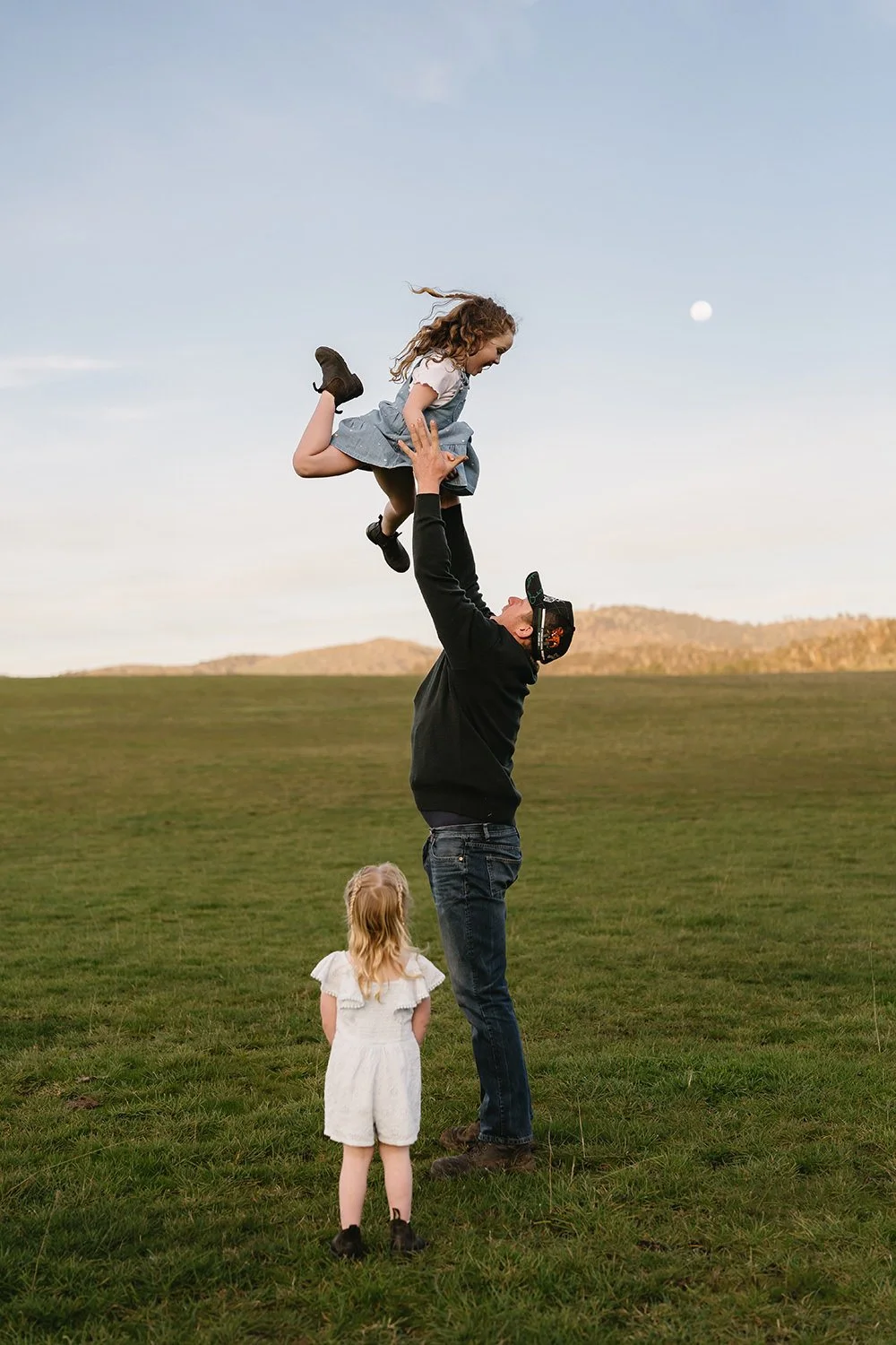 A man lifting a young girl into the air in an open grassy field during sunset, with two other children watching nearby and a mountain in the background.