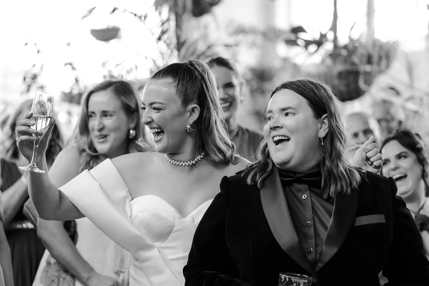 Group of cheerful women at a celebration, with one woman in a white dress holding a glass of champagne, all smiling and laughing.
