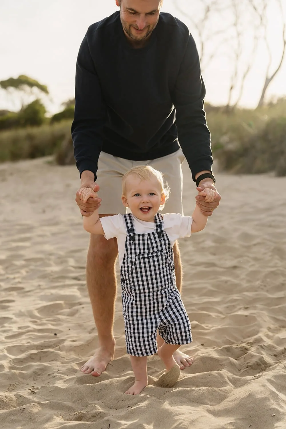 A man and a young child holding hands and smiling on a sandy beach with trees in the background, during sunset.
